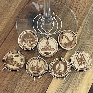 Puede incluir: Seis charms de copa de vino de madera con diferentes monumentos y nombres de ciudades. Los charms están unidos a anillos plateados. Los charms están sobre una superficie de madera.