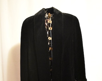 May include: A long, black velvet coat with a wide lapel collar and cuffed sleeves. The coat is displayed on a metal stand against a white backdrop. The coat's length extends to the ankles.