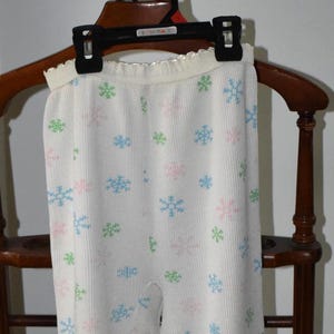 Op de afbeelding: Witte thermische pyjamabroek met een sneeuwvlokkenpatroon in roze, blauw en groen. De broek hangt aan een houten kledingstandaard. De manchetten en tailleband zijn wit.
