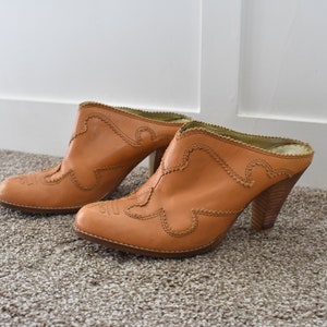 May include: A pair of brown leather heeled mules with a western-style design. The shoes have a pointed toe and a decorative stitch pattern.