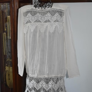 May include: A white blouse with long sleeves and detailed lace inserts. The blouse has lace panels across the chest and at the hem, creating a delicate design. The garment is displayed on a black wire mannequin.