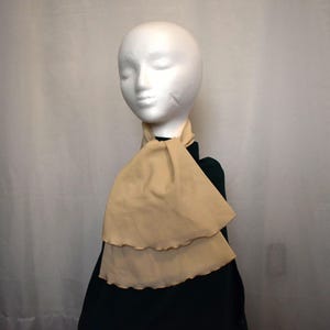 May include: A beige scarf with a layered design is draped around a mannequin's neck. The scarf has a soft, flowing appearance and is made of a lightweight fabric. The mannequin is wearing a dark green top.