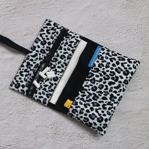 Könnte beinhalten: Eine schwarz-weiß-Leopardenmuster-Tasche mit schwarzem Griff. Die Tasche ist offen und zeigt ein schwarzes Innenfutter mit einem weißen und schwarzen Reißverschluss sowie mehrere weiße zylindrische Objekte. Ein kleines gelbes und blaues Objekt ist sichtbar.