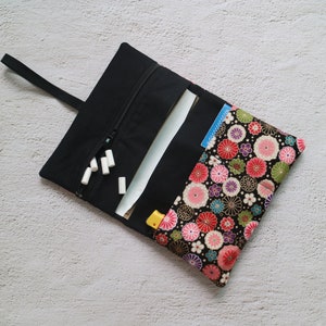 Könnte beinhalten: Schwarzes Stoff-Roll-Up-Etui mit floral gemusterter Klappe. Das Etui ist geöffnet und zeigt weiße Kreidestifte, Papiere und einen kleinen gelben Gegenstand. Das Blumenmuster umfasst rosa, rote und grüne Blumen auf schwarzem Hintergrund.