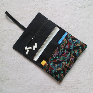 Könnte beinhalten: Eine schwarze Stofftasche mit Reißverschluss und einem bunten Drachenmuster als Futter. Die Tasche ist geöffnet und enthält weiße Kreidesticks.