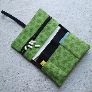 Könnte beinhalten: Grüne Stoff-Rolltasche mit schwarzem Reißverschluss und Riemen. Die Tasche ist geöffnet und zeigt ein schwarzes Innenfutter, weißes Papier und einen kleinen gelben Gegenstand. Mehrere weiße zylindrische Objekte sind sichtbar.