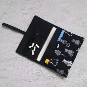 Könnte beinhalten: Schwarze Stoff-Rolltasche mit Reißverschlusstasche, weißer Papierhülle und schwarzem Riemen. Die Tasche ist mit weißen Katzensilhouetten auf schwarzem Hintergrund verziert. Mehrere weiße Kreidestifte und ein gelbes Feuerzeug sind sichtbar.