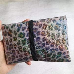 Könnte beinhalten: Eine rechteckige Clutch mit einem schimmernden Leopardenmuster in Silber-, Lila- und Grüntönen. Ein schwarzes Gummiband sichert die Clutch. Die Clutch wird in einer Hand gehalten.