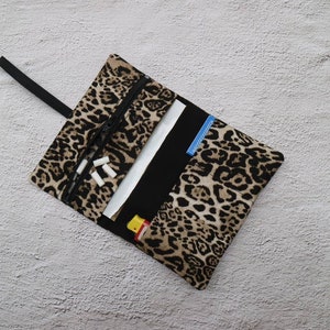 Könnte beinhalten: Eine aufrollbare Tasche mit Leopardenmuster, schwarzem Reißverschluss, schwarzem Riemen und schwarzem Innenraum. Die Tasche ist geöffnet und zeigt weißes Papier, einen blauen Akzent und kleine weiße Gegenstände. Die Tasche liegt auf einer hellgrauen Oberfläche.