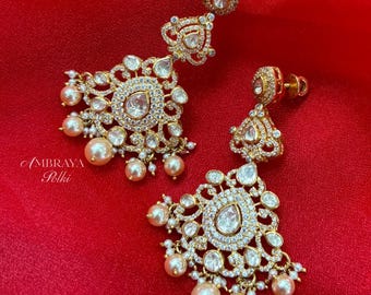 May include: Gold-toned earrings with a floral design, featuring clear gemstones and dangling pearls. The earrings have a central teardrop-shaped gemstone and are set against a red background. The text "AMBRAYA Polki" is visible.