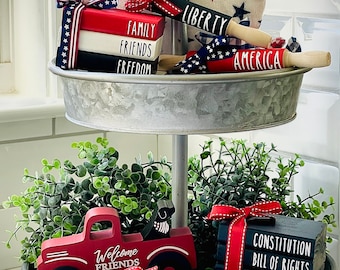Pode incluir: Uma bandeja de metal de dois níveis com um tema patriótico vermelho, branco e azul. O nível superior apresenta um rolo de madeira com a palavra "Liberty" nele, um rolo de madeira com a palavra "America" nele e uma pilha de blocos de madeira com as palavras "Family", "Friends" e "Freedom" neles. O nível inferior apresenta um caminhão de brinquedo vermelho com as palavras "Welcome Friends" nele, um rolo de madeira com a palavra "Patriot" nele e uma pilha de blocos de madeira com as palavras "Constitution", "Bill of Rights" e "Freedom" neles.