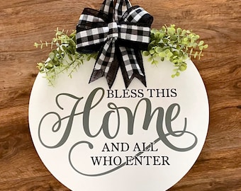 May include: A white round wooden door hanger with the text "Bless this home and all who enter" in grey lettering. The door hanger is decorated with a black and white checked bow and a sprig of green foliage.