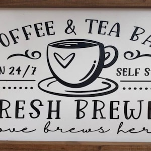 May include: A white sign with black text and a black and white illustration of a coffee cup. The sign reads "Coffee & Tea Bar, Open 24/7, Self Serve, Fresh Brewed, Love brews here."