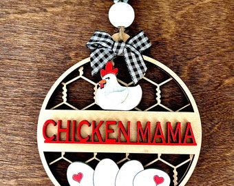May include: A wooden round sign with a chicken and eggs design. The sign has a red and white checkered bow, a white chicken with a red comb, and the words "Chicken Mama" in red letters. The sign is surrounded by a wire mesh design.