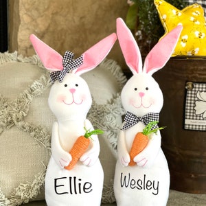 May include: Two white plush Easter bunnies with pink ears and black and white checkered bows. The bunnies are holding carrots and have the names "Ellie" and "Wesley" embroidered on their chests.