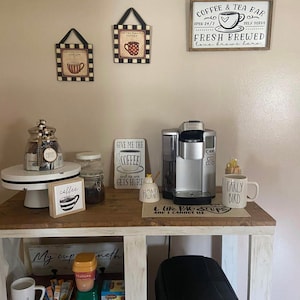 May include: A white wooden coffee bar with a coffee maker, coffee mugs, and a variety of coffee and tea supplies. The bar has a sign that says "Coffee & Tea Bar" and "Fresh Brewed Love Brews Here".