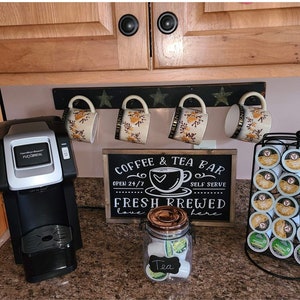 May include: A black coffee maker with a silver trim sits on a countertop next to a sign that reads "Coffee & Tea Bar Open 24/7 Self Serve Fresh Brewed Love Here". There are four mugs hanging on a rack above the sign, and a clear glass jar filled with tea bags labeled "Tea".