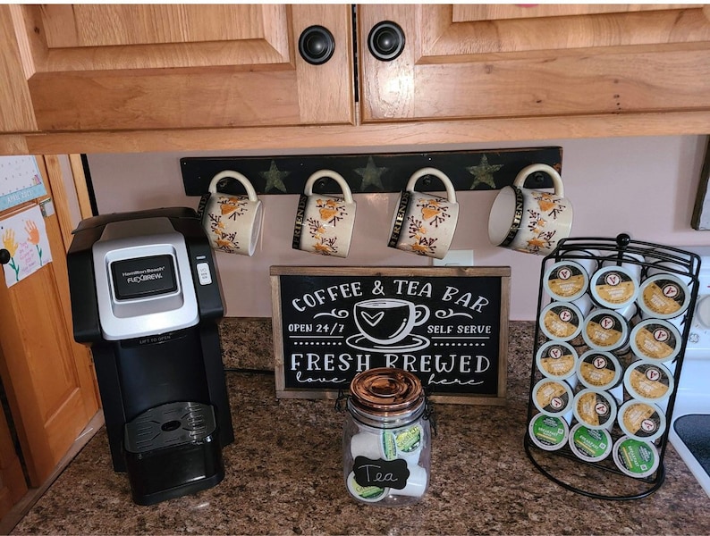 May include: A black coffee maker with a silver trim sits on a countertop next to a sign that reads "Coffee & Tea Bar Open 24/7 Self Serve Fresh Brewed Love Here". There are four mugs hanging on a rack above the sign, and a clear glass jar filled with tea bags labeled "Tea".