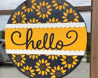 Può includere: Un appendiporta rotondo in legno con sfondo nero e una fascia gialla con la parola "hello" in lettere nere. Lo sfondo è decorato con un motivo di girasoli neri.