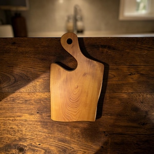 May include: A light brown wooden cutting board with a natural wood grain pattern, featuring a curved handle with a circular hole at the top. The board has a smooth, flat surface and an irregular, organic shape with rounded edges and a warm tone.