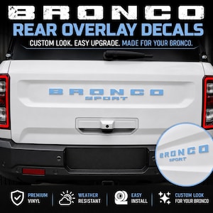 May include: Light blue Bronco Sport rear overlay decals applied to the white tailgate of a vehicle. The text BRONCO SPORT is centered on the tailgate, with a circular inset showing a close-up view of the light blue lettering on white paint.