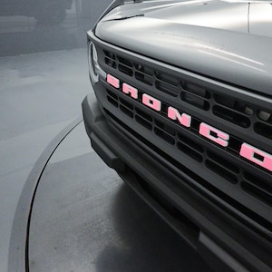May include: Close-up of a gray vehicle's front grill with the word "BRONCO" in pink letters. The grill is black and has a horizontal slat design. The vehicle's hood is visible above the grill.