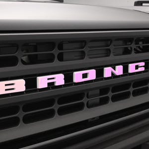 May include: Close-up of a black car grille with the word "BRONCO" in pink letters. The grille has a series of horizontal and vertical bars. The car's hood and headlight are visible in the background.