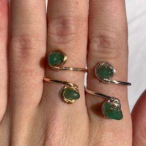 May include: Two wire-wrapped rings with green gemstones. One ring is gold-toned, the other is silver-toned. The rings are on a finger, showcasing the jewelry's design and color. The gemstones are a light green color.