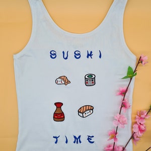 Pode incluir: Camisola branca de alças com o texto "SUSHI TIME" bordado a azul. A camisola apresenta quatro imagens bordadas com tema de sushi: uma peça de sushi, um rolo de sushi, uma garrafa de molho de soja e uma peça de nigiri sushi.