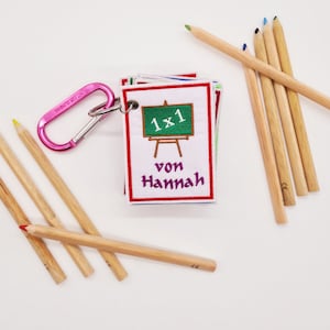 Peut inclure: Un ensemble d'étiquettes en tissu colorées avec un tableau vert et le texte "1x1 von Hannah" sur fond blanc. Les étiquettes sont attachées à un mousqueton rose et sont entourées de crayons en bois.