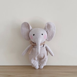 May include: A plush gray elephant toy wearing a brown and white gingham overall and a brown knitted scarf.