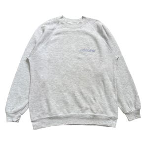 Op de afbeelding: Een lichtgrijs gemêleerd sweatshirt met ronde hals, lange mouwen en geribbelde boorden bij de kraag, mouwen en zoom. Op de borst staat een klein, lichtblauw geborduurd Adidas-logo met de merknaam en het klaverbladsymbool in het midden.