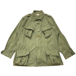 May include: An olive green long-sleeve field jacket featuring a pointed collar, shoulder epaulets, and four large front pockets with buttoned flaps. The garment has a central button-down closure and long sleeves with adjustable buttoned cuffs.