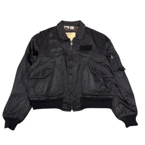 May include: Black bomber jacket with a zip-up front, two front pockets, and a small pocket on the left sleeve. The jacket has a ribbed collar, cuffs, and hem. A small patch is visible on the chest.
