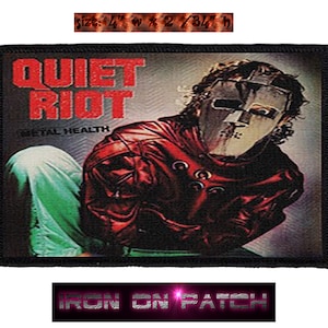 May include: A rectangular patch featuring the band Quiet Riot. The patch displays the band's name in red block letters above an image of a person wearing a mask and red jacket. The patch measures 10.16 cm wide by 6.99 cm high.