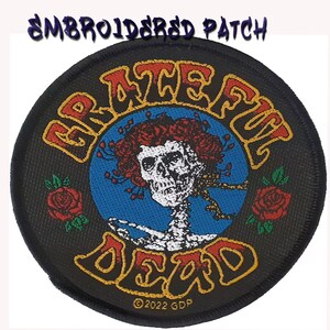Peut inclure: Un écusson brodé circulaire sur fond noir avec le texte "GRATEFUL DEAD" en orange et rouge. Le centre présente un crâne blanc entouré de roses rouges sur un cercle bleu, avec deux roses rouges supplémentaires sur les côtés.
