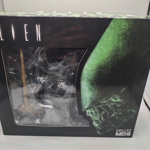 Puede incluir: Una caja negra con la palabra "ALIEN" en letras blancas. La caja presenta una imagen verde y negra de una criatura alienígena. La figura es visible a través de la ventana de plástico transparente. La caja también tiene el texto "DELUXE MDS".