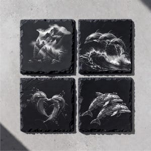 May include: Set of four square black slate coasters. Each coaster features a different black and white dolphin design. One shows a mountain scene, another dolphins jumping, a third with dolphins forming a heart, and the last with steampunk dolphins.