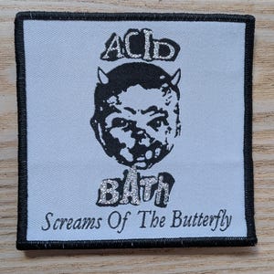 Peut inclure: Écusson carré avec une bordure noire et un fond bleu clair. Le patch présente les mots "ACID BATH" au-dessus d'une illustration noire d'un visage avec des cornes. Le texte "Screams Of The Butterfly" est en dessous.