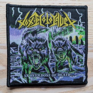 Op de afbeelding: Vierkante patch met een zwarte rand. Het heeft een paarse en groene illustratie van twee grommende wolven. De patch heeft de tekst "An Overdose of Death..." en de bandnaam in geel.