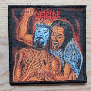 May include: Square patch with the word "DEICIDE" in red above a graphic illustration. The illustration depicts two figures, one with a blue mask and the other with a grimacing face. The patch has a black border.
