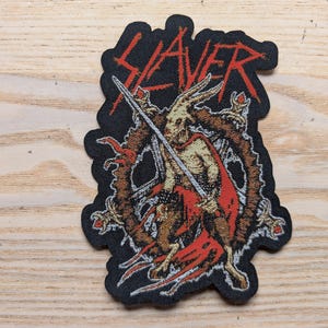May include: Black embroidered patch with the word "Slayer" in white lettering. The patch features a red and white illustration of a creature with a sword.