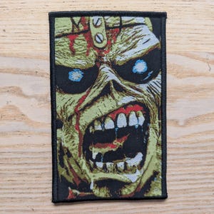 Può includere: Una toppa rettangolare con bordo nero presenta un'illustrazione dettagliata di un volto simile a uno zombie. Il volto è reso in tonalità di verde, giallo e rosso, con occhi blu e bocca aperta.