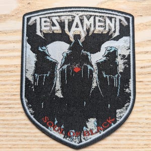 Puede incluir: Parche bordado con el nombre de la banda "TESTAMENT" en letras plateadas sobre una ilustración oscura de figuras encapuchadas. La frase "SOUL OF BLACK" está arqueada en la parte inferior. El parche tiene forma de escudo con borde negro.
