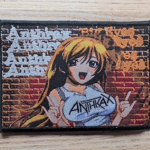 Könnte beinhalten: Gestickter Aufnäher mit einem Anime-Charakter mit blonden Haaren, der ein blaues Oberteil mit dem Bandnamen "ANTHRAX" trägt. Der Hintergrund ist eine Backsteinmauer mit dem Bandnamen in stilisiertem Text. Der Aufnäher hat einen schwarzen Rand.