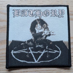 Può includere: Toppa quadrata con bordo nero con la scritta "BATHORY" in lettere gotiche argentate. Sotto, un'immagine in bianco e nero di una persona e un pentagramma.