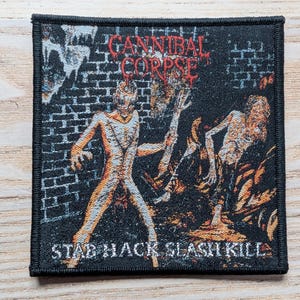 May include: Square black patch with the words "CANNIBAL CORPSE" in red, above a graphic of two figures in a brick wall setting. The text "STAB HACK SLASH KILL" is below the graphic. The figures are in shades of orange and white.