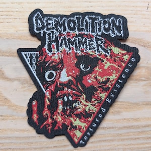 May include: A black patch featuring the words "DEMOLITION HAMMER" in white, above a red and yellow graphic of a face. The words "Tortured Existence" are printed along the right side of the patch.