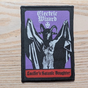 Pode incluir: Remendo retangular com borda preta e fundo roxo. Apresenta uma ilustração preta de uma figura com chifres e braços estendidos. O texto "Electric Wizard" está no topo e "Lucifer's Satanic Daughter" na parte inferior.