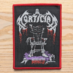 May include: Black rectangular patch with a red border featuring the word "Mortician" in white gothic lettering above a skeletal chair design. The phrase "Hacked Up For Barbecue" is printed at the bottom.
